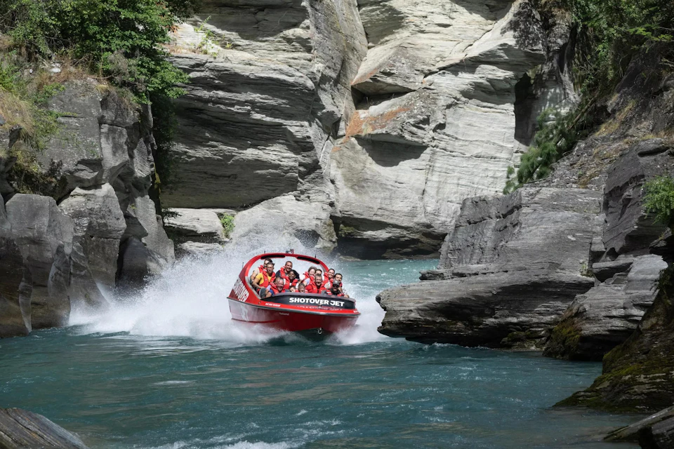A red jet boat with passengers moves swiftly through a narrow canyon with turquoise water and rocky cliffs.