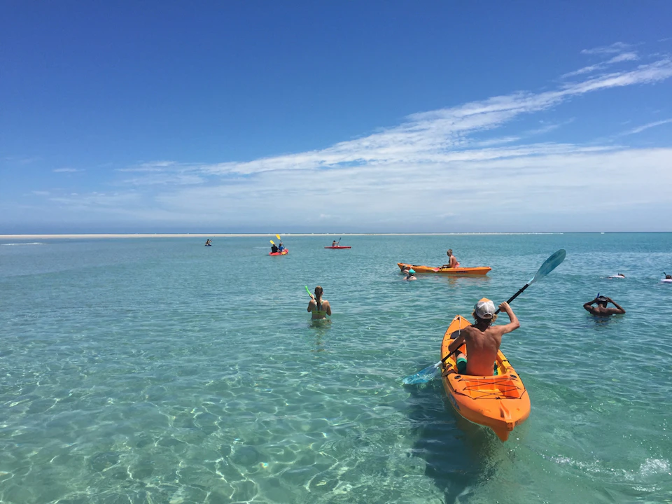 People kayaking and swimming in clear, shallow ocean water under a bright blue sky with scattered clouds.