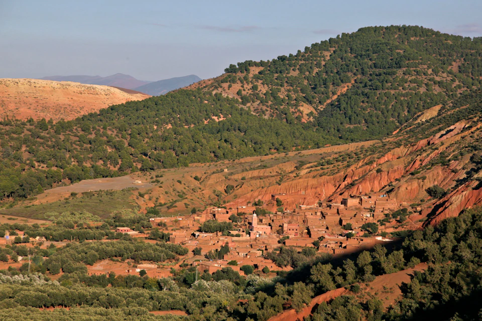A village with traditional houses nestled in a valley surrounded by green hills and trees under a blue sky.