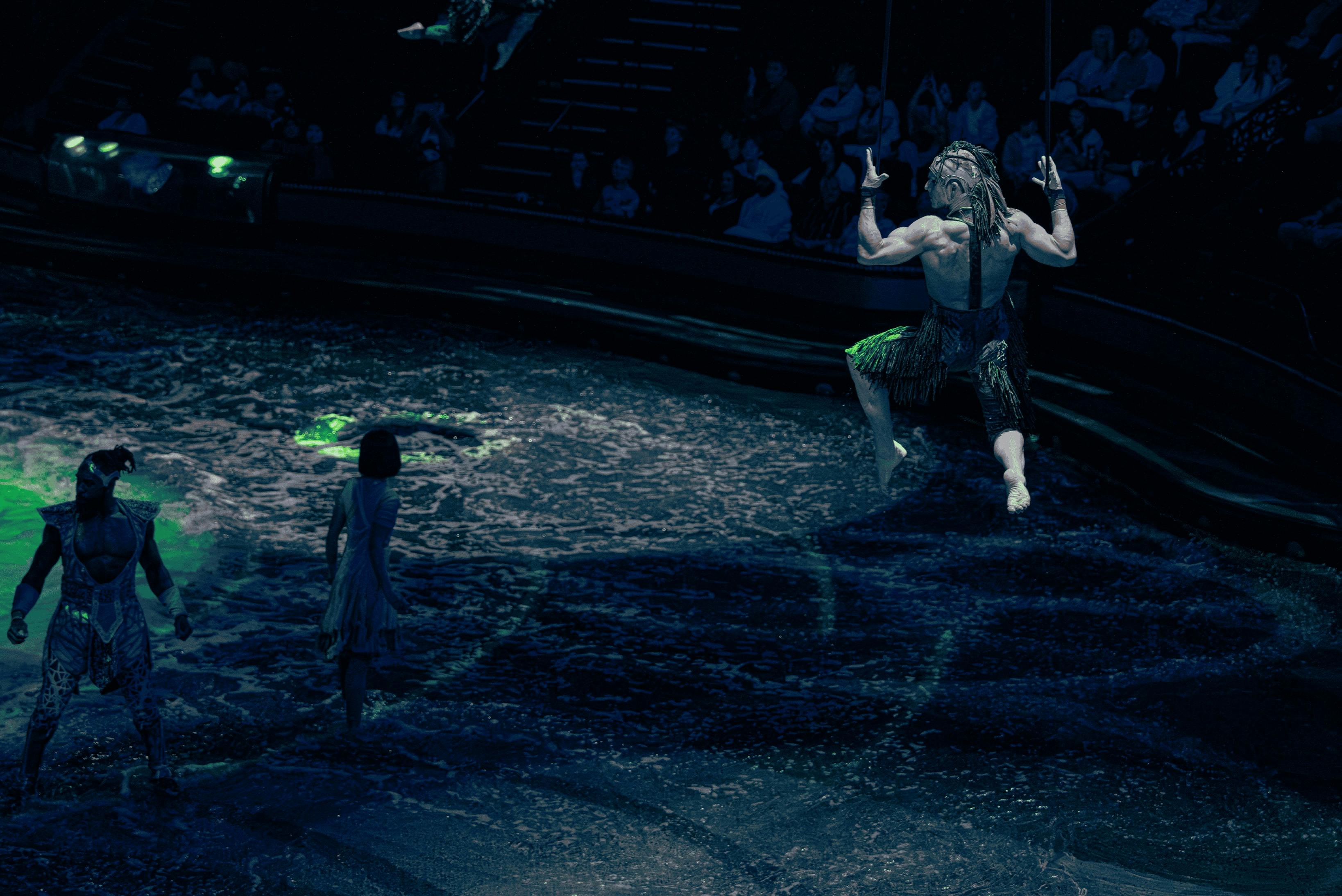 Een artiest in groene kleding hangt in de lucht terwijl het publiek toekijkt, met een kind dat op een met water bedekt podium staat.