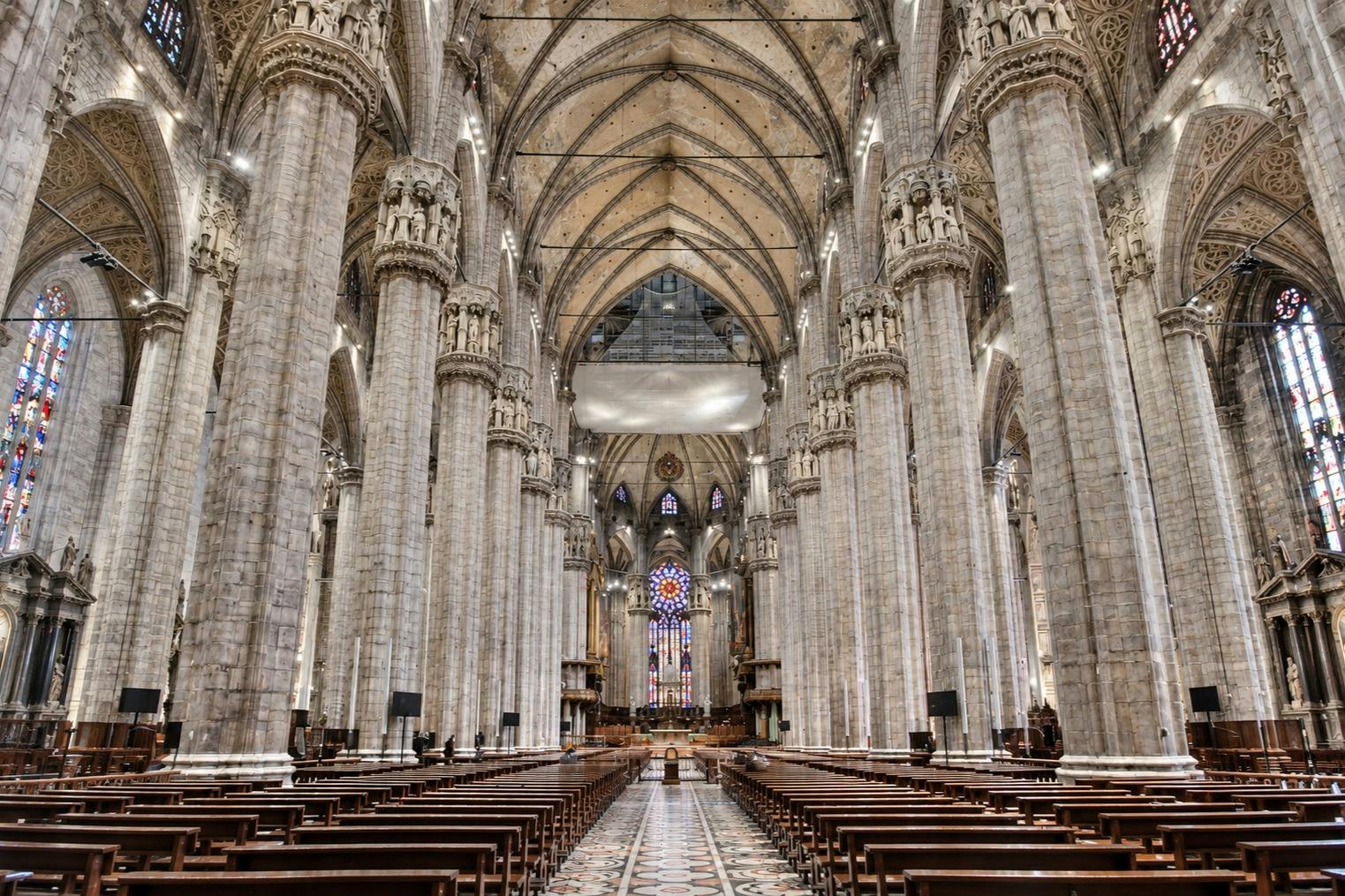 Interior view of a grand cathedral with tall stone columns, high vaulted ceilings, and a stained glass window at the end.