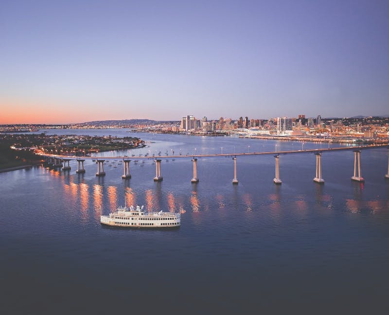 City skyline at dusk with a large boat on the water and a bridge connecting two landmasses.