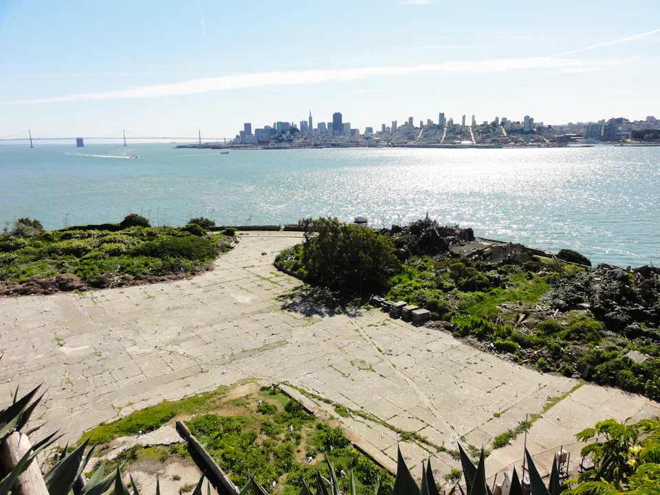 View of a waterfront cityscape across a bay from a stone-paved area with greenery in the foreground.