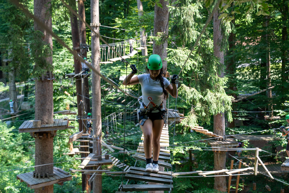 A person wearing safety gear and a green helmet walks on a rope bridge in a forest adventure park.