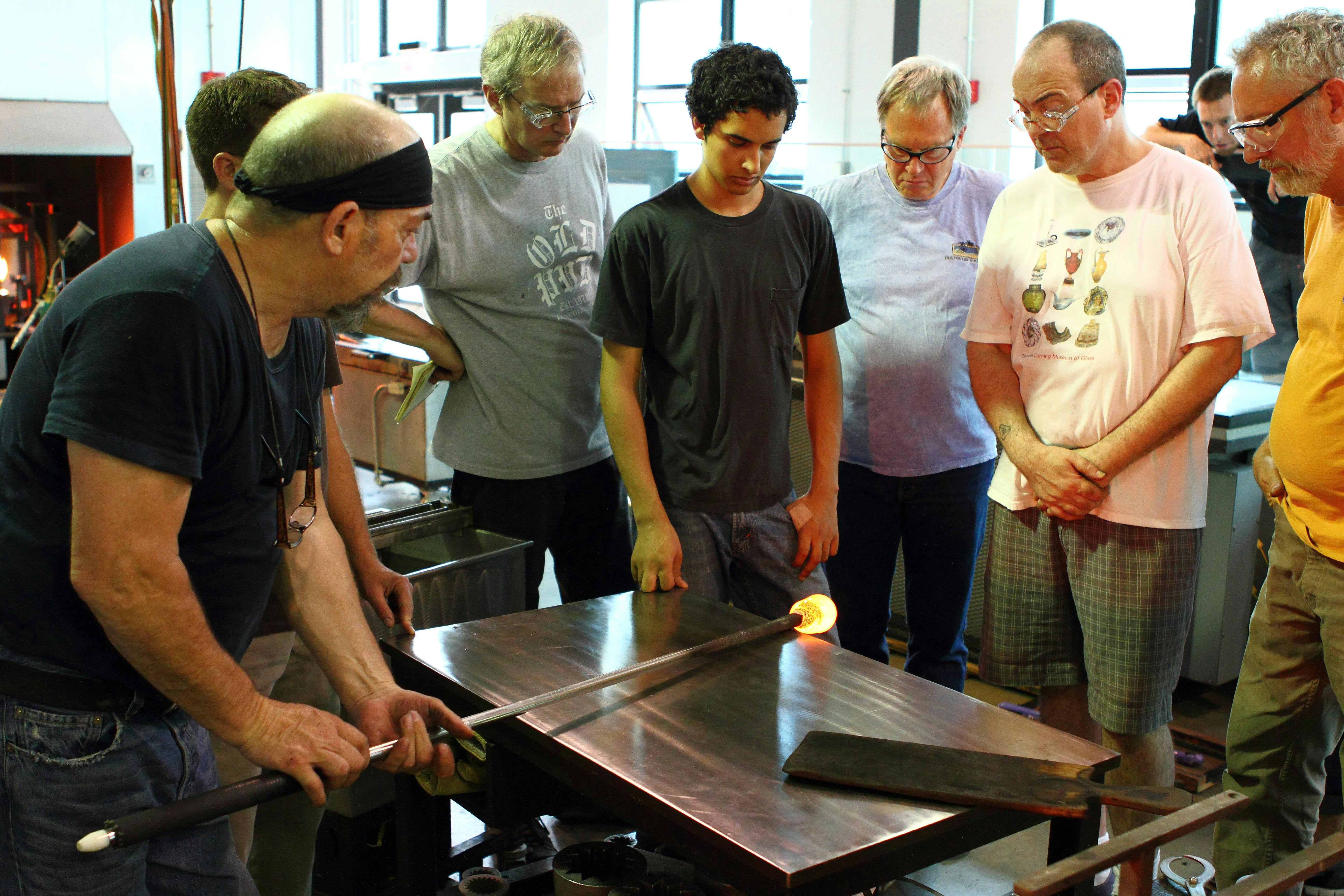 A group of people observing a person shaping molten glass on a rod on a workbench in a workshop setting.