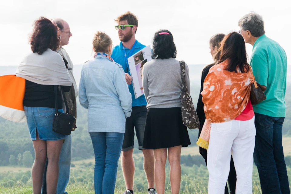 A man in a blue shirt and sunglasses speaks to a group of people outdoors near a grassy hill and a scenic background.