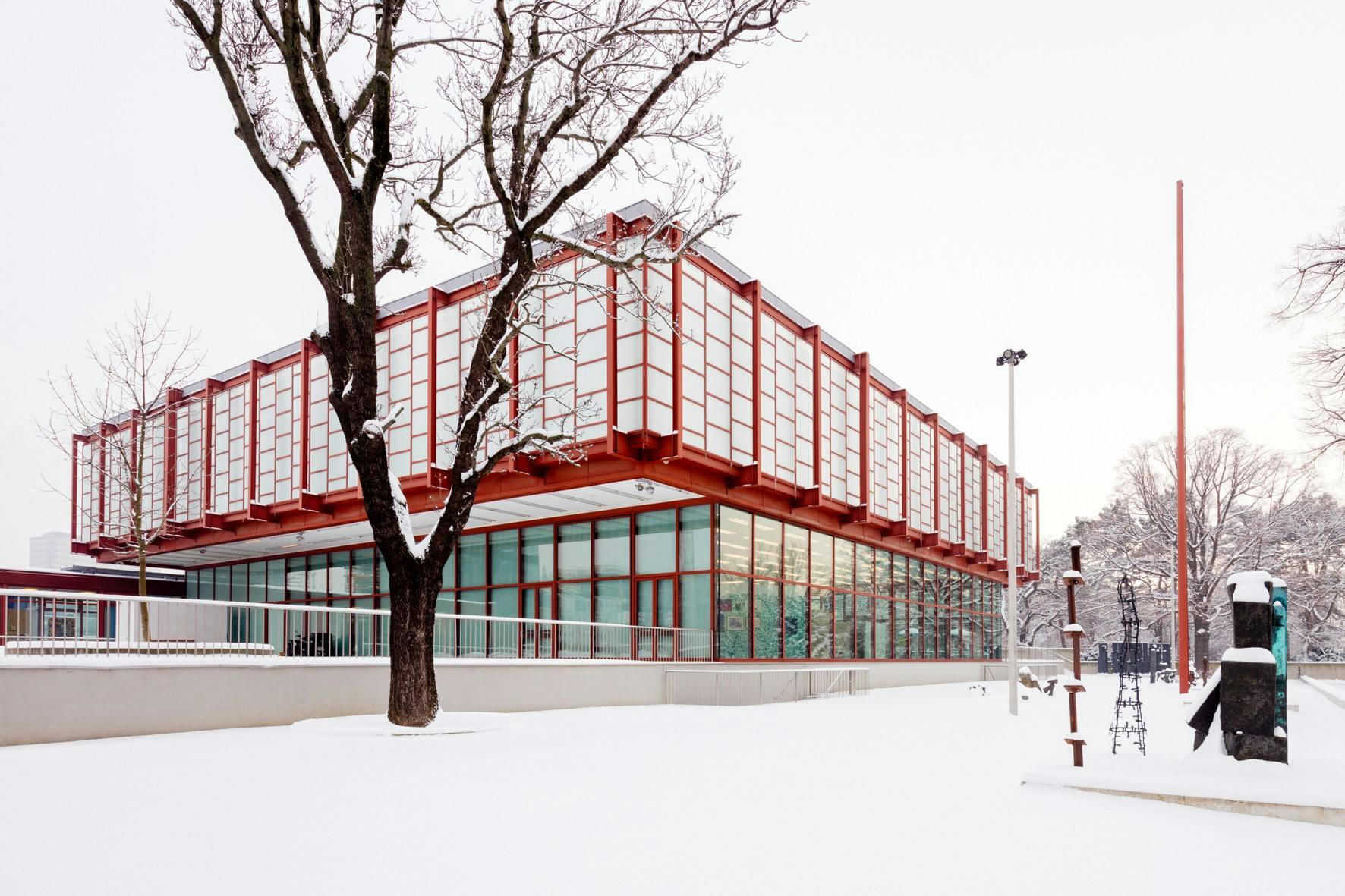 Un bâtiment moderne avec une charpente rouge et de grandes fenêtres dans un paysage enneigé, avec des arbres dénudés et des sculptures abstraites à proximité.