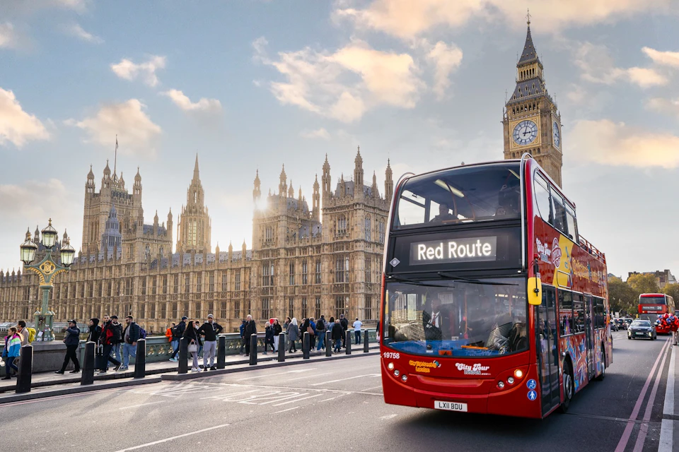 A red double-decker bus labeled 'Red Route' drives on a bridge near the Houses of Parliament and Big Ben in London.