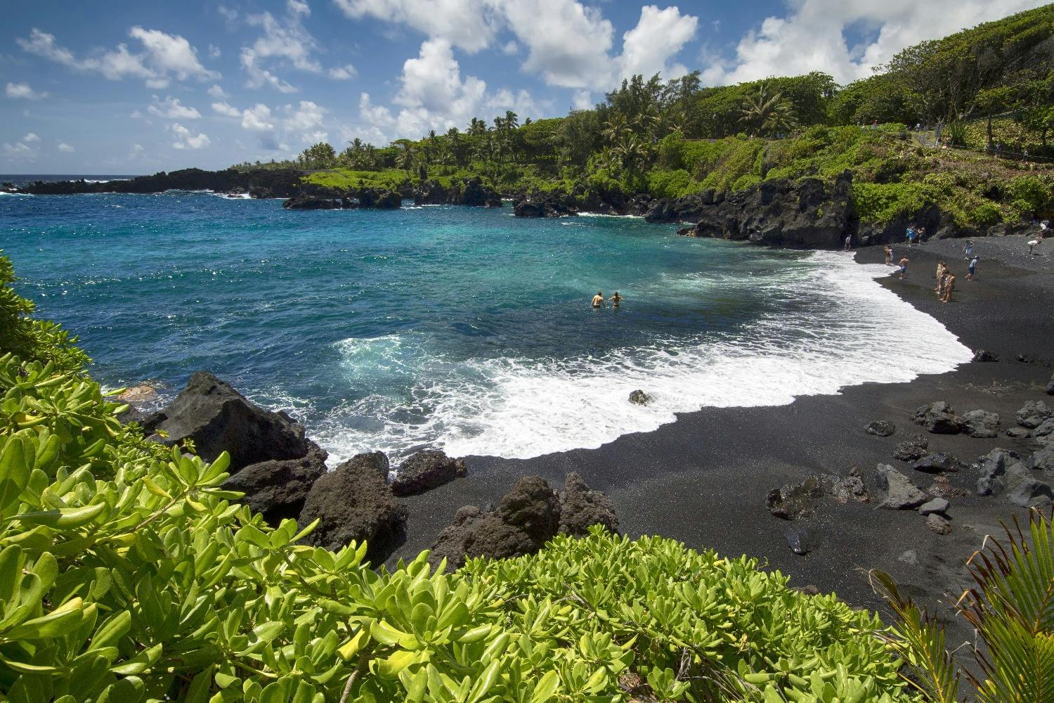 A tropical beach with black sand, lush greenery, and clear blue waters. Two people swim near the shore under a partly cloudy sky.