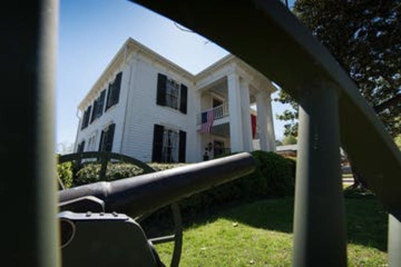 Wit huis van twee verdiepingen met zwarte luiken, geflankeerd door een Amerikaanse vlag, gezien vanachter een oud kanon op een zonnige dag.