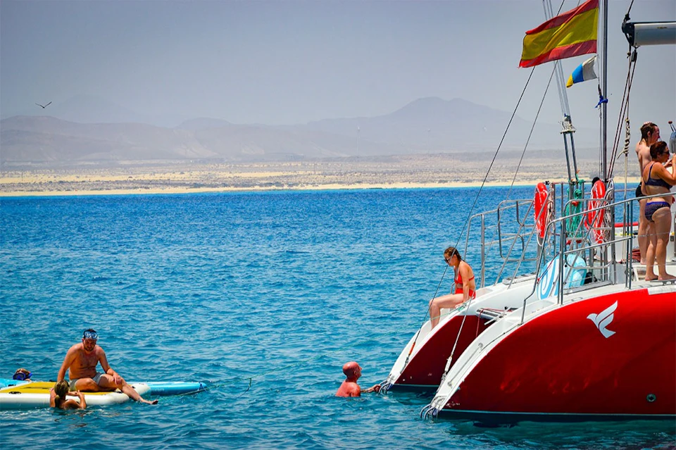 People enjoying the water near a boat with red hulls and a flag, against a backdrop of mountains and clear blue sea.