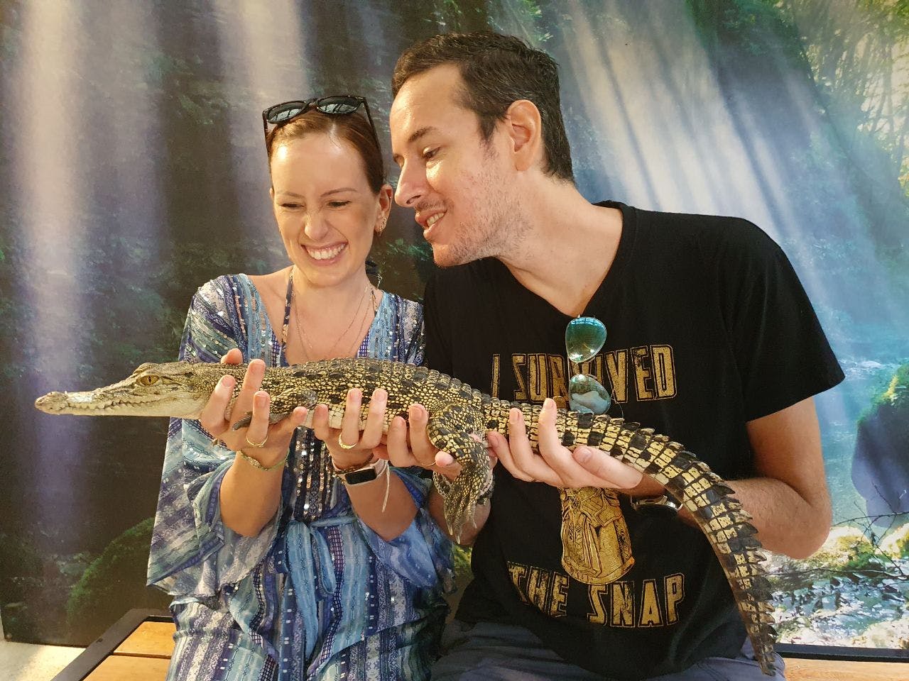 两个人微笑着，手里拿着一条小鳄鱼，后面是一片森林。