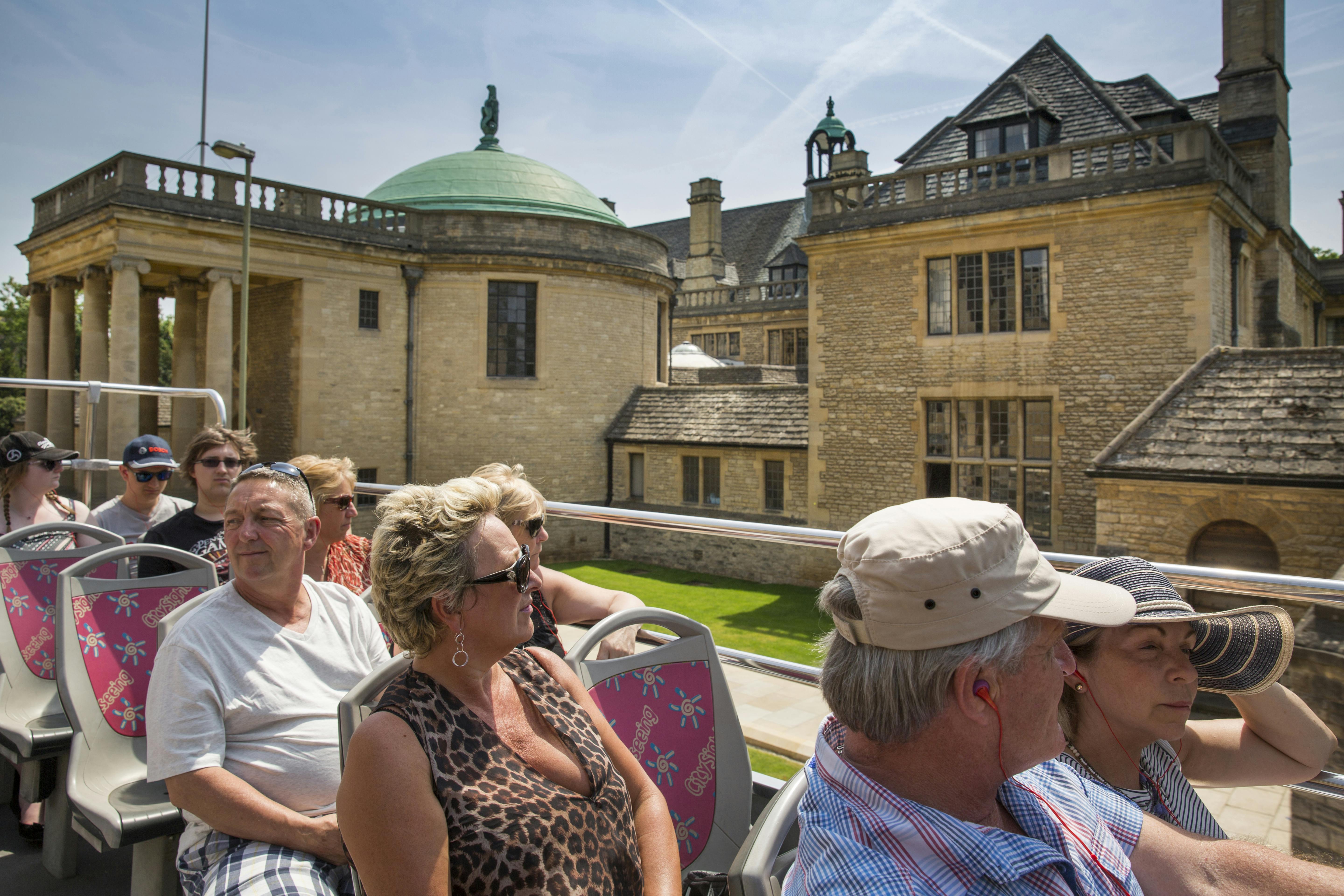 Tourists on an open-top bus ride, seated and viewing historic stone buildings with a grassy courtyard in the background.