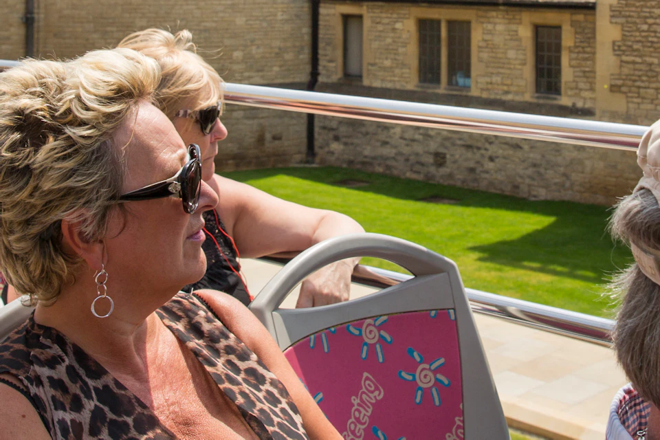 Tourists on an open-top bus ride, seated and viewing historic stone buildings with a grassy courtyard in the background.