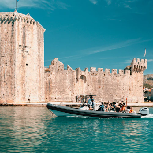 En båt med människor som rider i lugnt vatten nära ett historiskt stentorn och fästningsmur, med byggnader i bakgrunden.