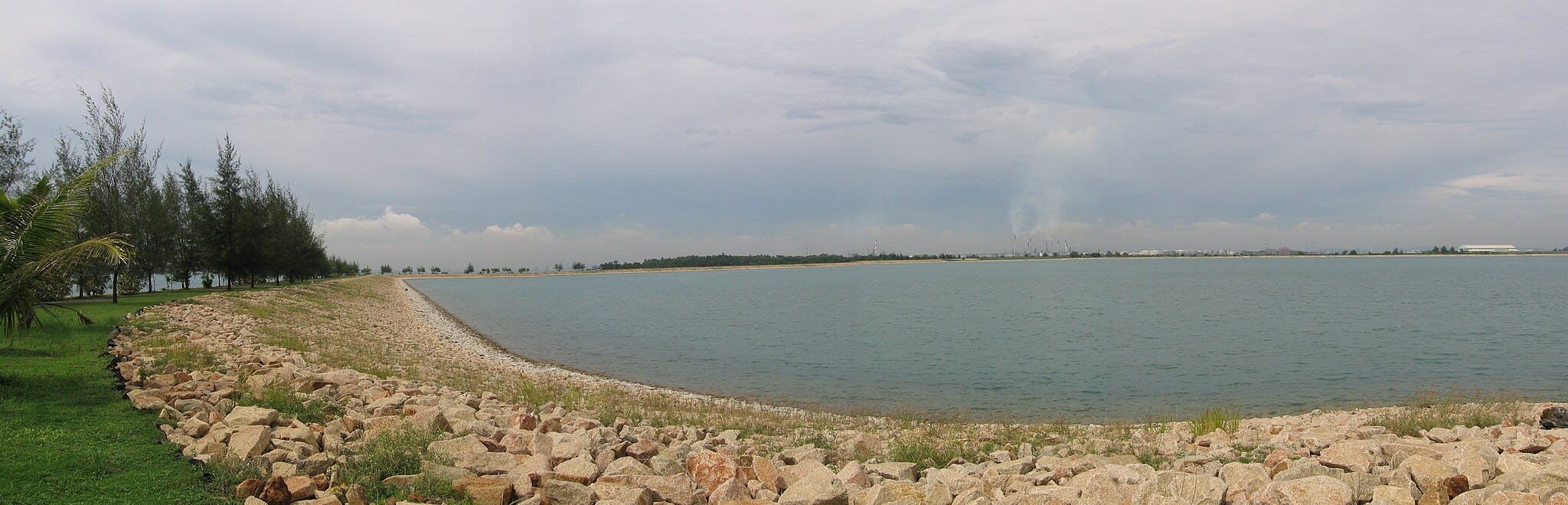 A rocky shoreline curves around a calm body of water, flanked by trees and greenery under a cloudy sky. Buildings in the distance.