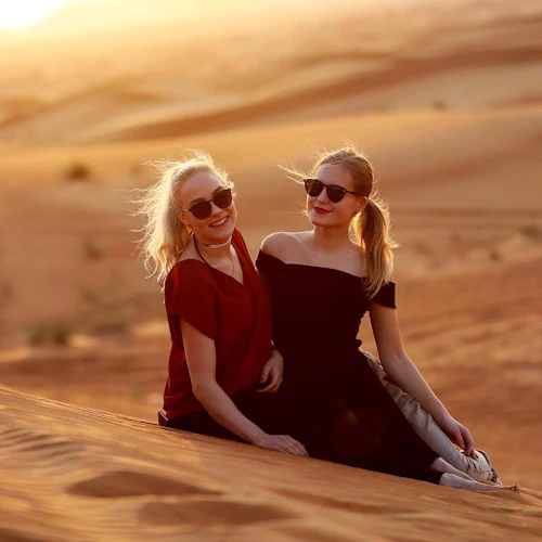 Zwei Frauen mit Sonnenbrillen sitzen auf einer Sanddüne bei einem warmen, goldenen Sonnenuntergang in einer Wüstenlandschaft.