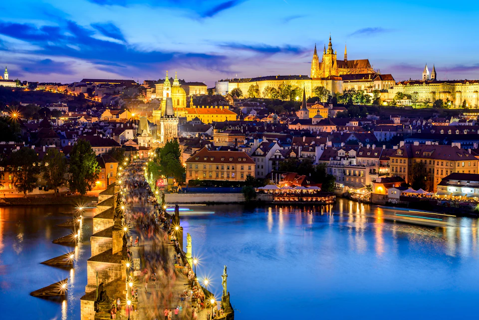 Illuminated cityscape at dusk featuring a busy stone bridge over a river, lit buildings, and a cathedral in the background.