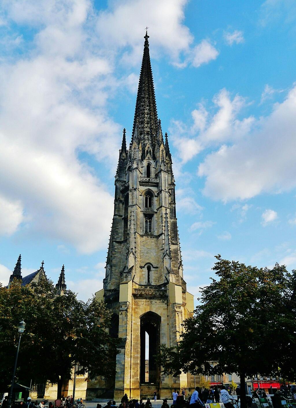 木々に囲まれ、一部曇り空の下で前景を歩いている人々に囲まれた、尖塔のある背の高いゴシック様式の石造りの教会塔。