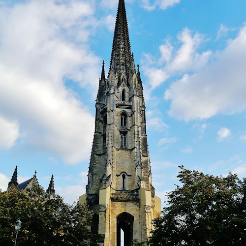 Ett högt stentorn i gotisk stil med en spiral, omgivet av träd och människor som går i förgrunden under en delvis molnig himmel.