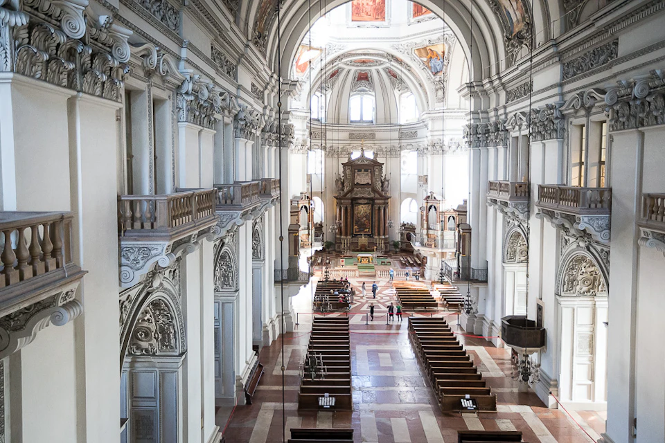 A spacious cathedral with ornate architecture, high arched ceilings, wooden pews, and a few visitors walking near the altar.