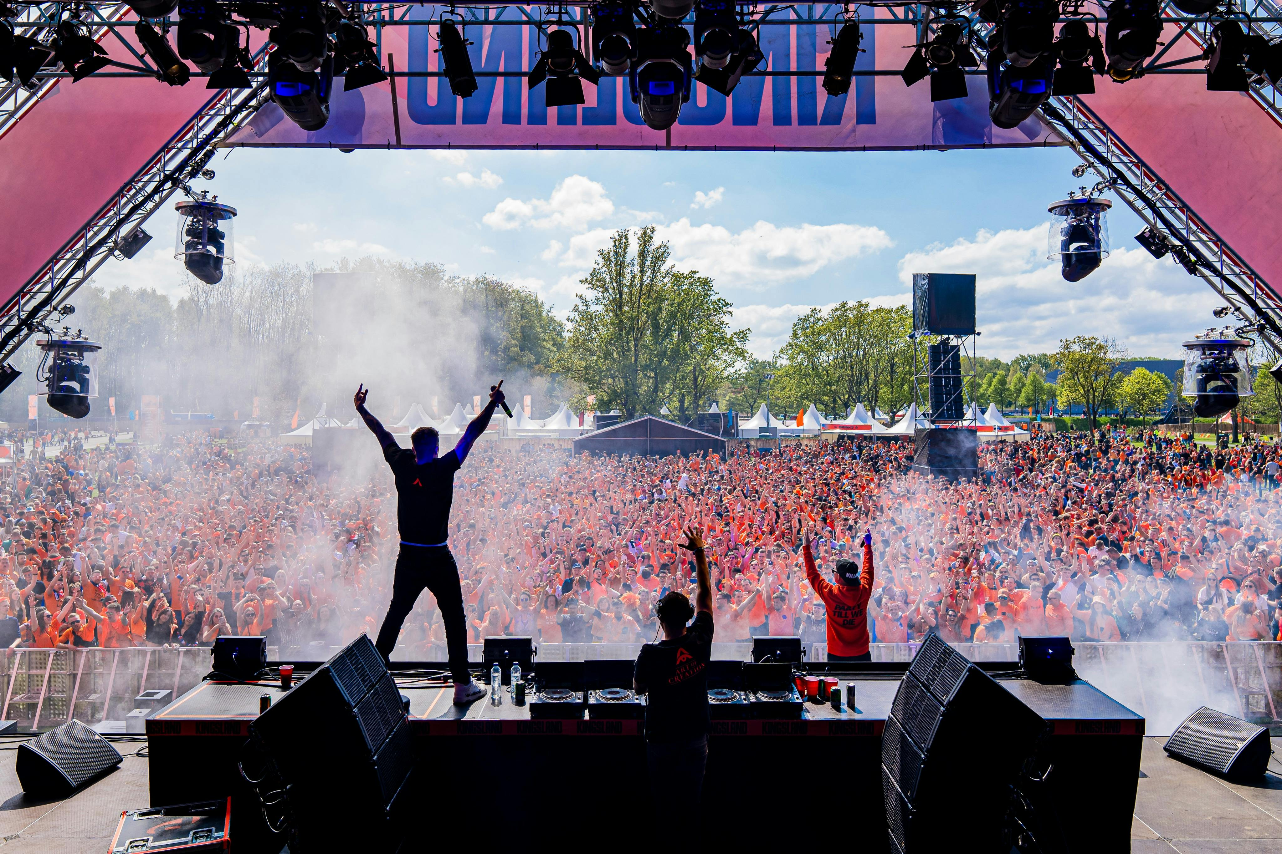 En stor publik jublar när två artister står på scenen, en med armarna lyfta, mitt bland rök och belysningsutrustning, utomhus under en ljus himmel.