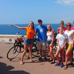 Grupp om sju personer som poserar med cyklar vid havet en solig dag; stadens silhuett och havet i bakgrunden.