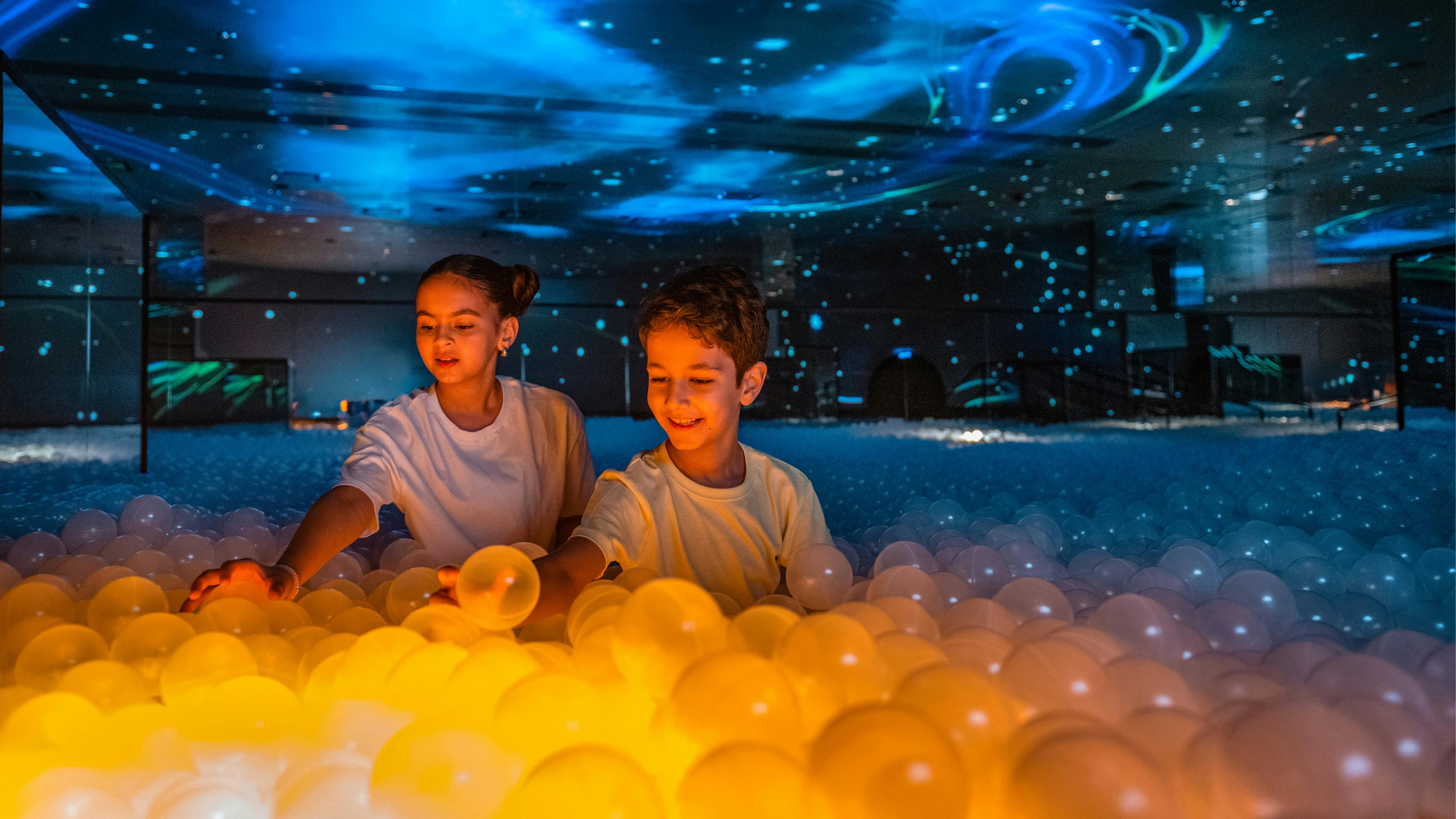 Δύο παιδιά παίζουν με φωτιζόμενες μπάλες σε ένα σκοτεινό δωμάτιο με μπλε και πορτοκαλί φωτισμό.