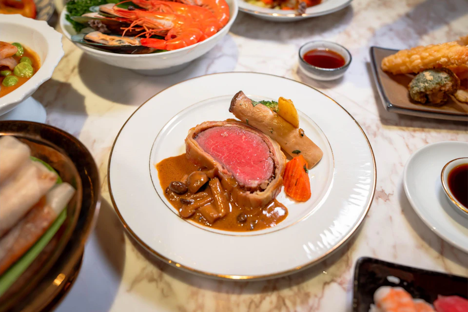 A plated dish featuring beef Wellington, vegetables, and sauce, with a bowl of shrimp and sauce in the background.