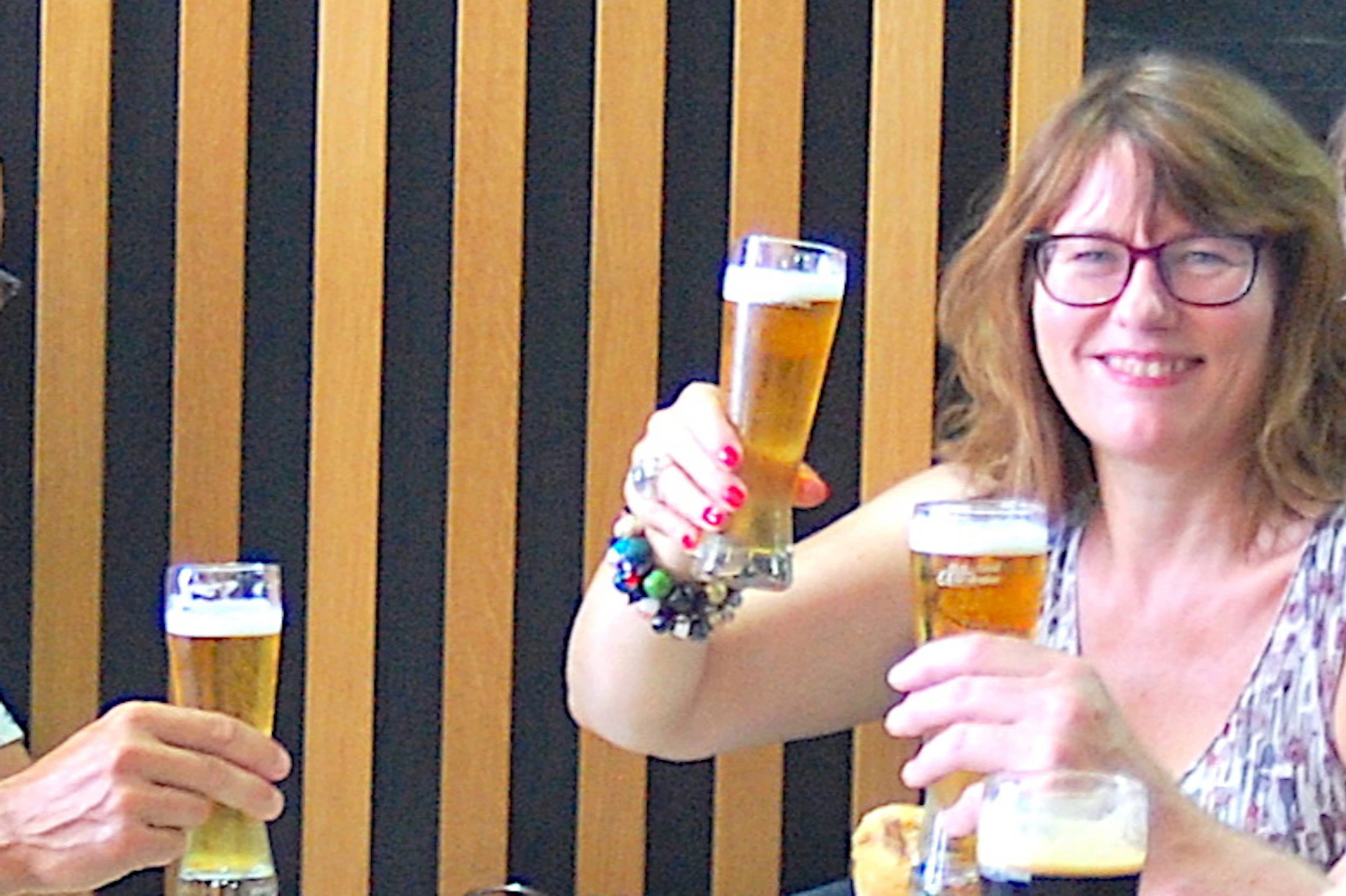 A group of six people smiles and raises glasses of beer at a table, enjoying burgers in a restaurant with a modern decor.