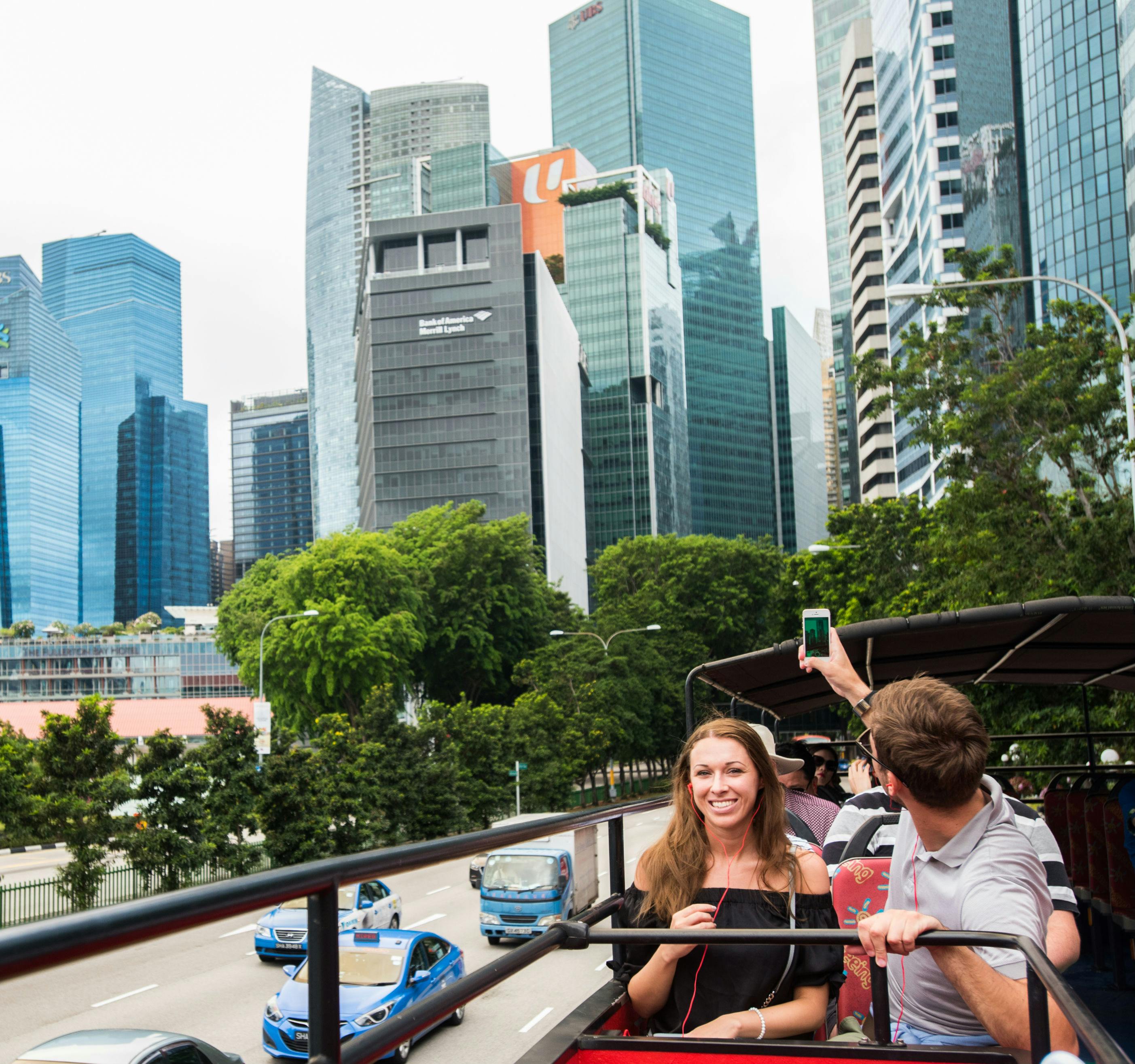 人们坐在敞篷巴士上，后面有摩天大楼和绿地。 一名男子用智能手机拍照。