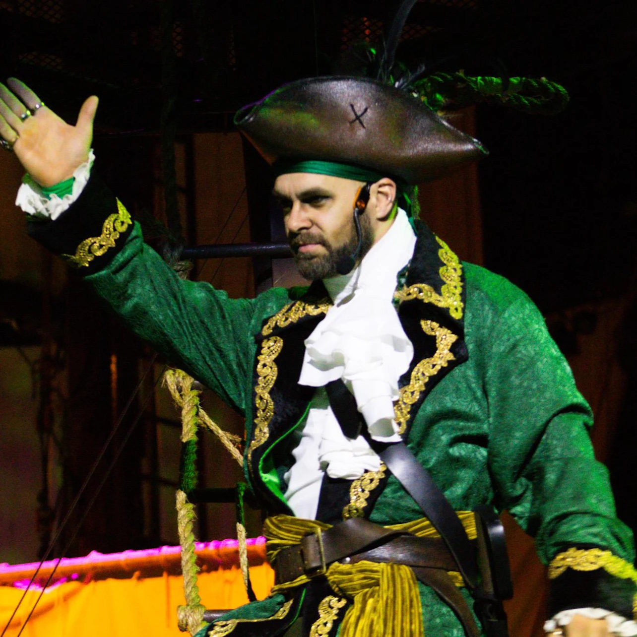 Man in a pirate costume with a green coat, black hat, and gold trim, raising one hand in a theatrical pose.