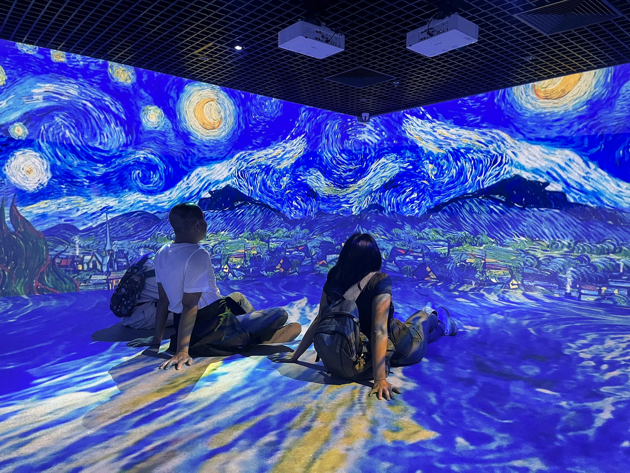 Deux personnes sont assises sur le sol, éclairé par une projection du tableau « La Nuit étoilée » de Van Gogh, dans une salle dédiée aux expériences immersives.