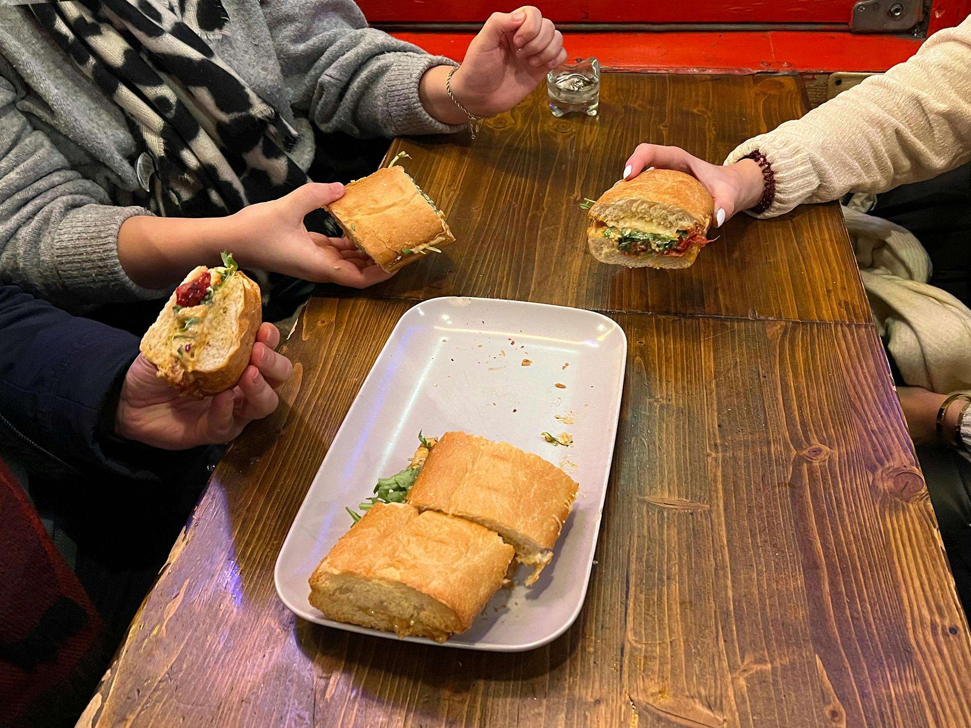 Τρία άτομα κρατούν σάντουιτς πάνω από ένα ξύλινο τραπέζι με ένα δίσκο που περιέχει κομμάτια σάντουιτς και ένα μικρό ποτήρι κοντά.