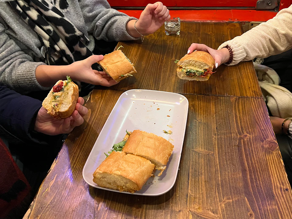 Three people holding sandwiches over a wooden table with a tray containing sandwich pieces and a small glass nearby.