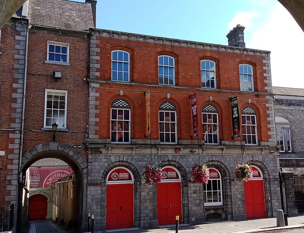 A red-brick building with arched windows, banners, and red doors. Hanging flower baskets decorate the front. A narrow alley runs through an archway to the left.