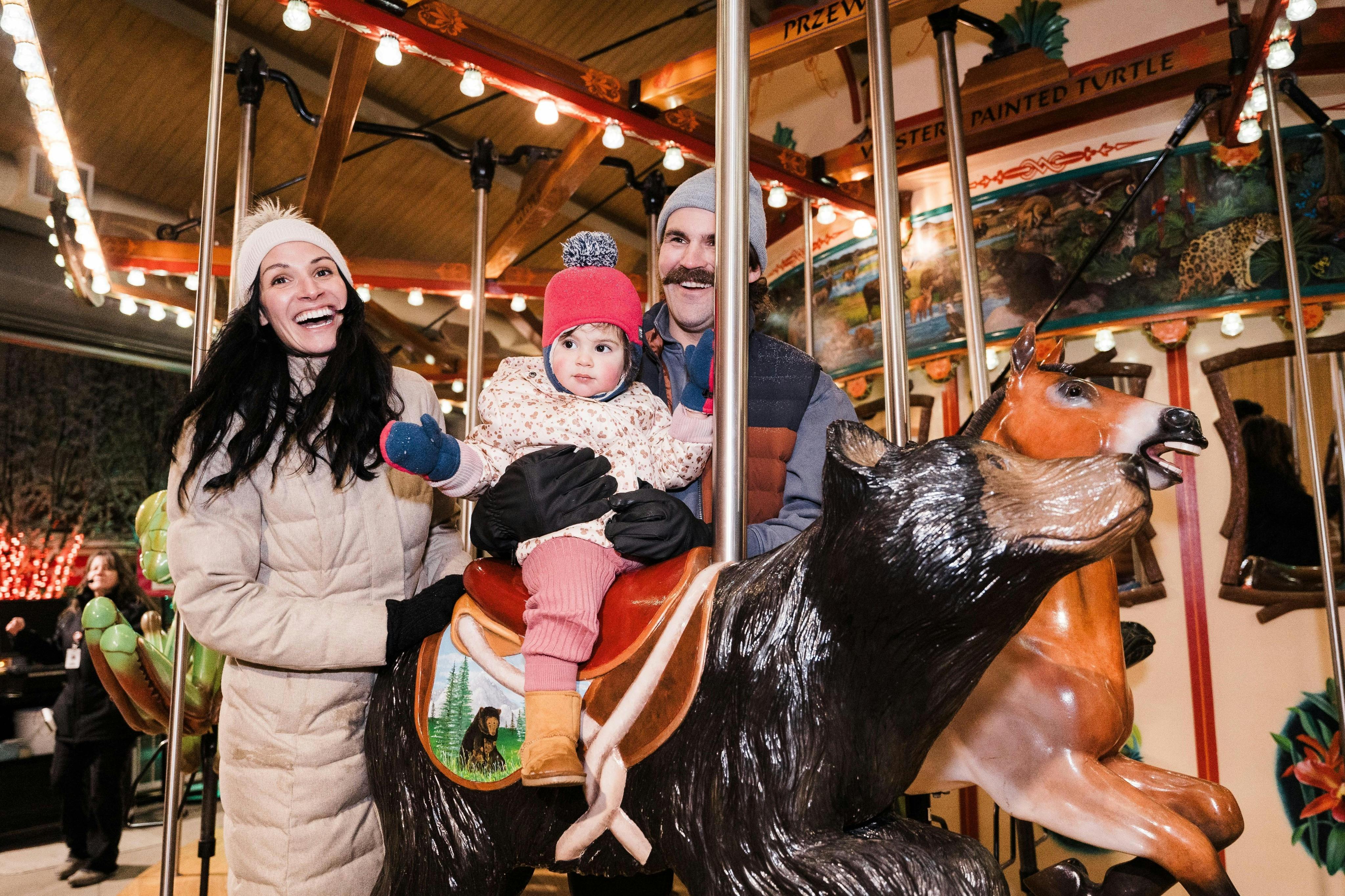 회전목마를 탄 두 명의 성인과 한 명의 어린이. 아이는 곰 위에 앉아 빨간 모자를 쓰고 있습니다. 어른들은 웃으며 겨울 복장에 묶여 있습니다.