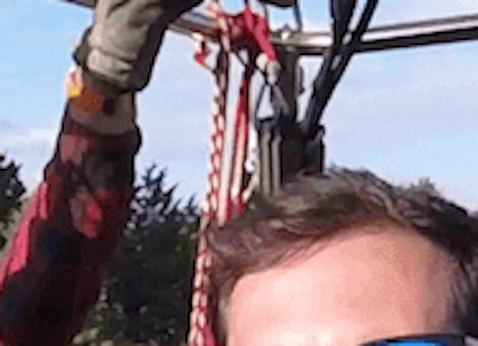 Three people smile and wave inside a hot air balloon basket, with another balloon visible in the background.