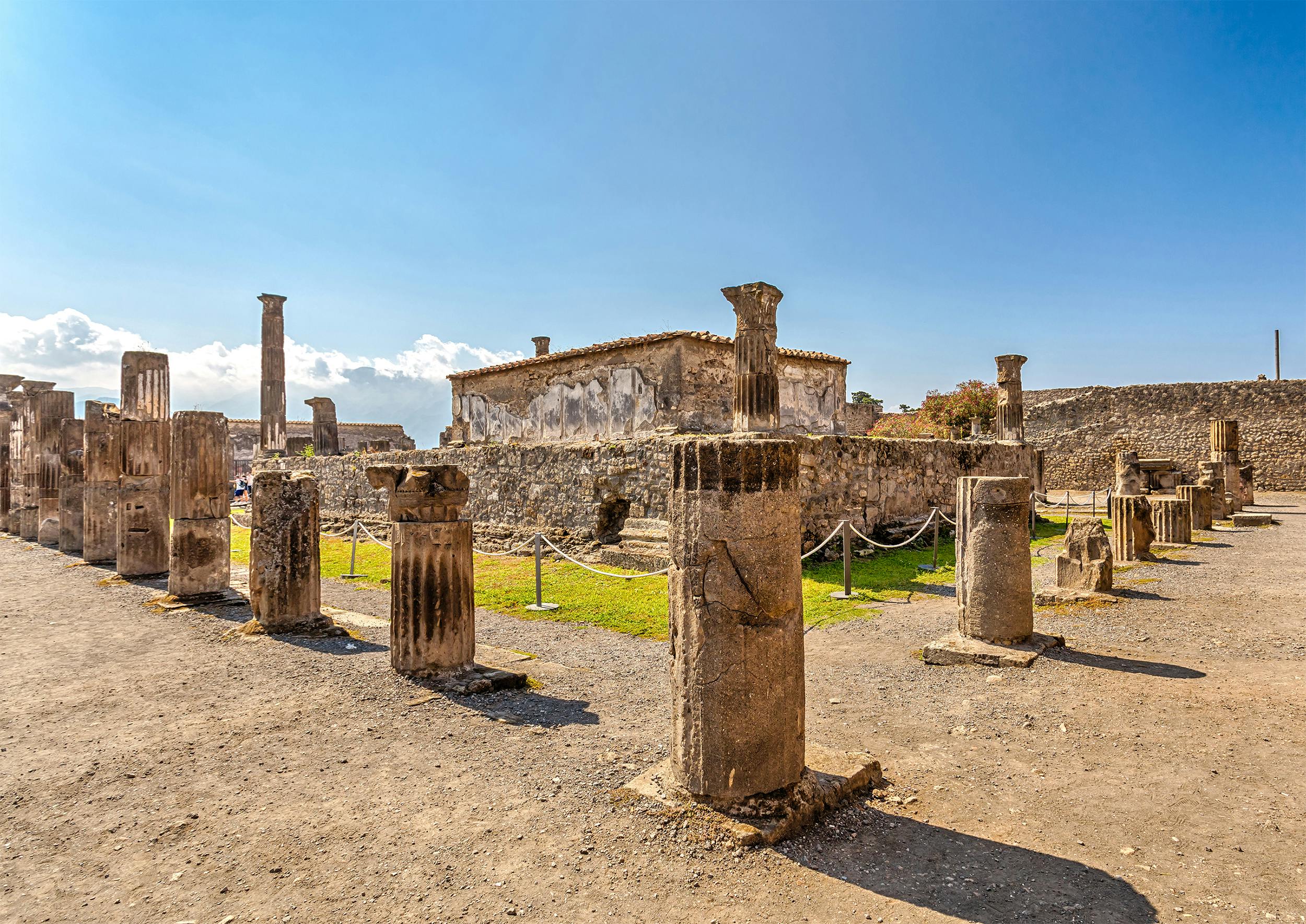 Ancient stone columns and ruins at an archaeological site under a clear blue sky. A building with weathered walls in the background.