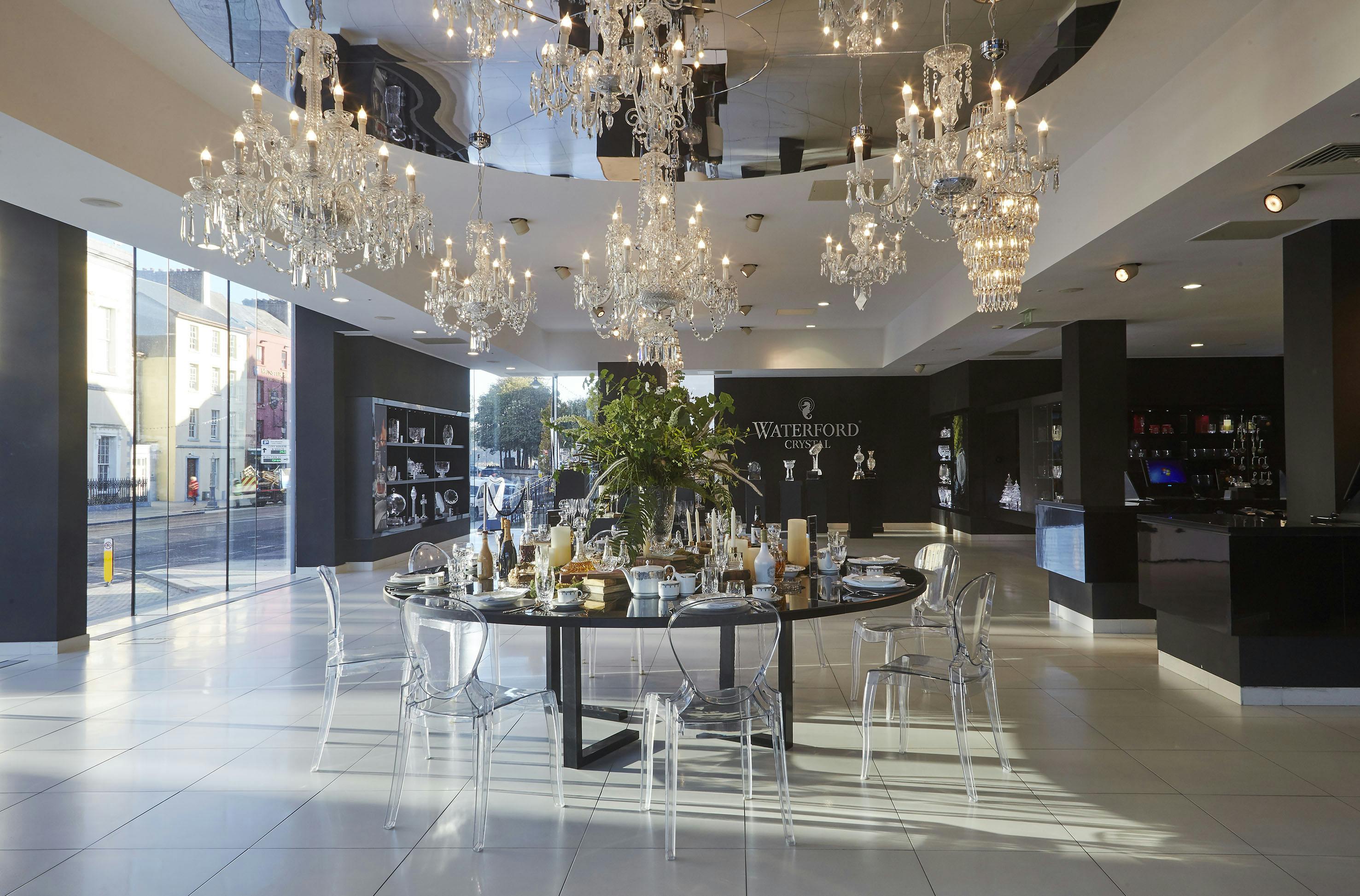 一间摆放着枝形吊灯的陈列室，摆放着圆形餐桌，四周环绕着透明的椅子。 墙上有沃特福德标志。
