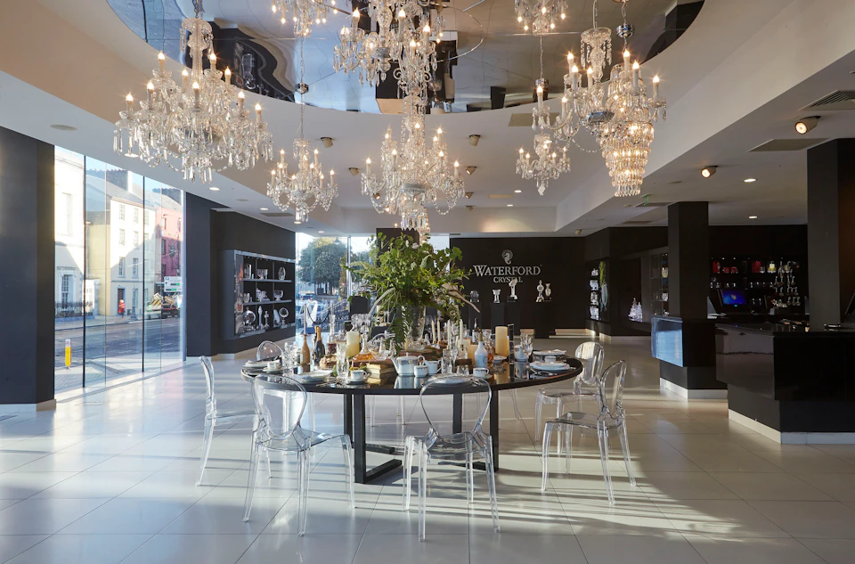 A showroom with chandeliers hanging over a circular table set for dining, surrounded by transparent chairs. Waterford logo on the wall.