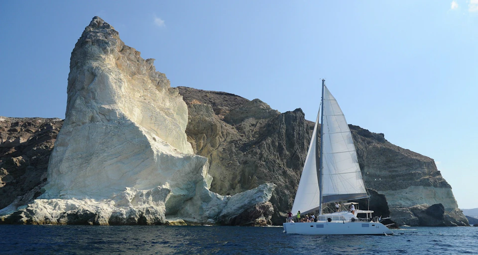 A sailboat with white sails is on the water near a rocky coastline under a clear blue sky.