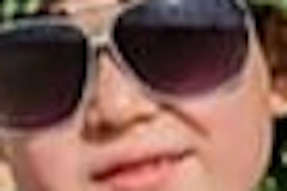 A child wearing sunglasses and a green headscarf poses with finger guns in front of a pyramid under a partly cloudy sky.