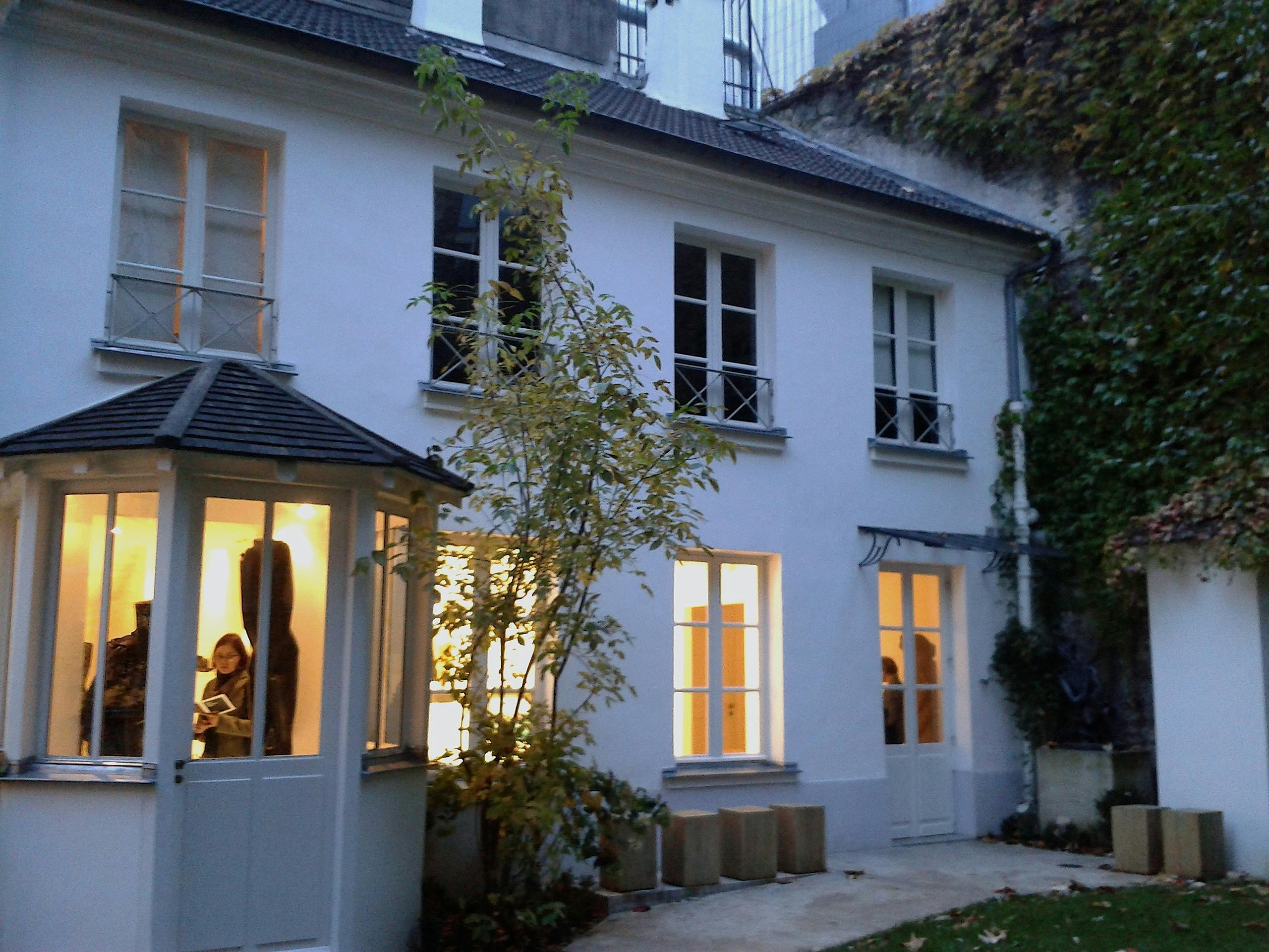 Διώροφο λευκό σπίτι με φωτισμένα παράθυρα. Οι άνθρωποι είναι ορατοί μέσα από τα μεγάλα παράθυρα. Ένα μικρό δέντρο στέκεται στον κήπο.