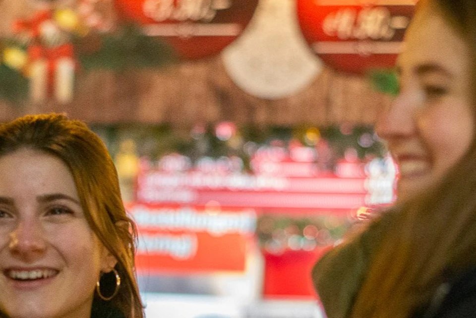 Two smiling individuals in warm clothing holding mugs, with festive decorations and signs in the background.