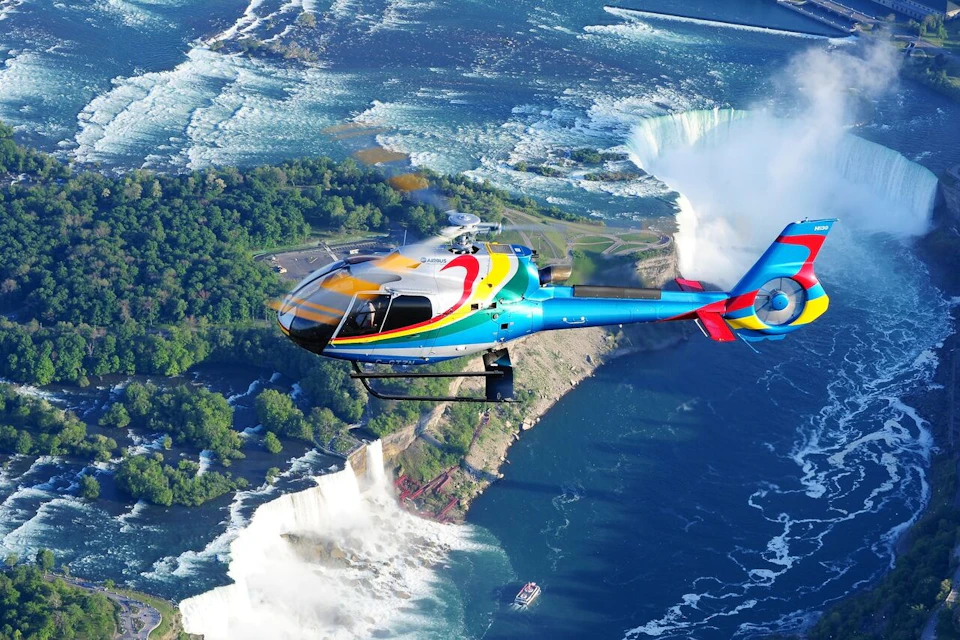 A colorful helicopter flies above a misty waterfall with lush green trees and turbulent water below.