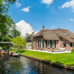 Ett tegelhus med halmtak vid en kanal; människor i båtar, en träbro och frodig grönska under en blå himmel.