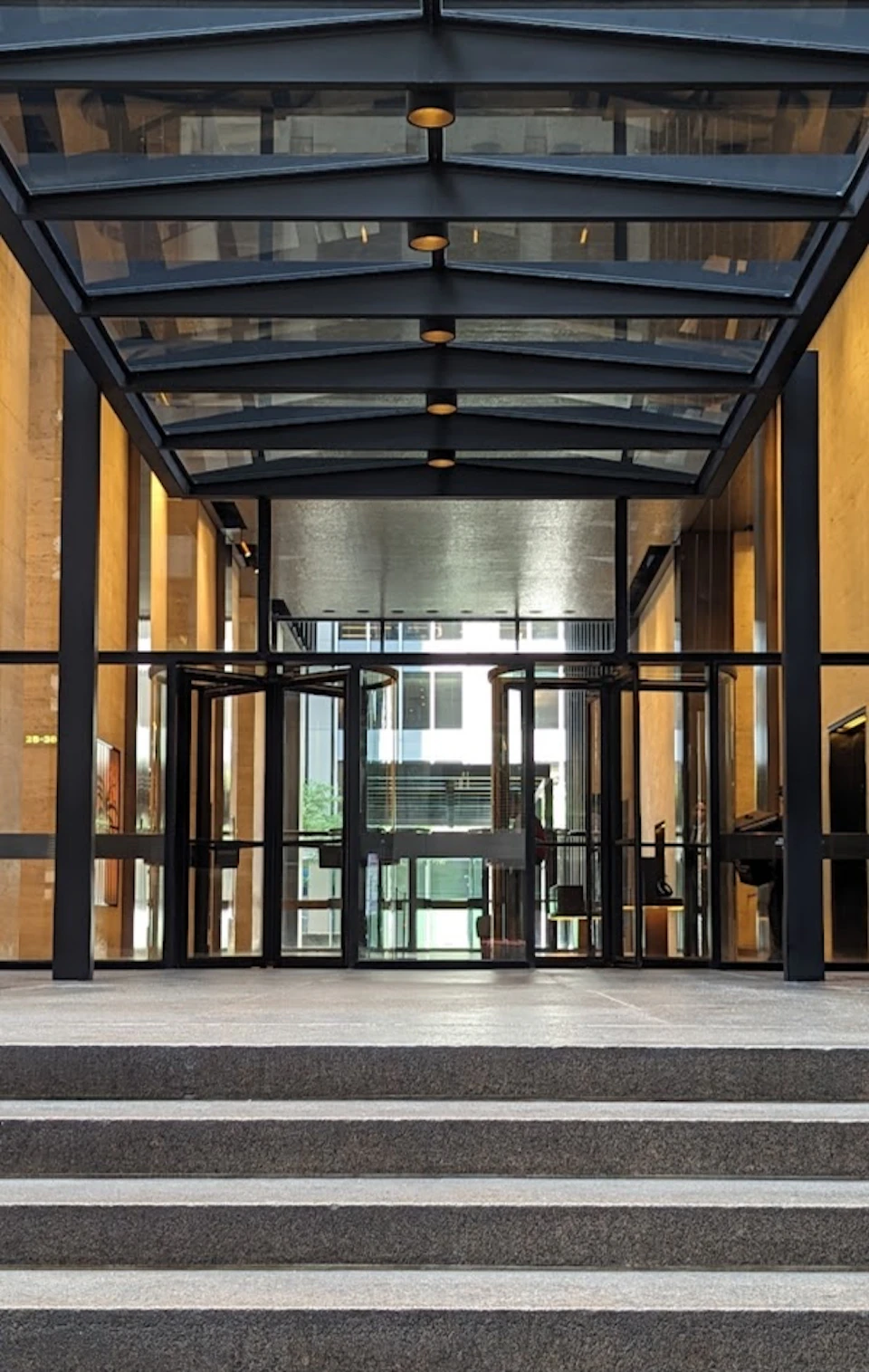 A glass entrance to a building with revolving doors, framed by a metal and glass canopy and illuminated interior.