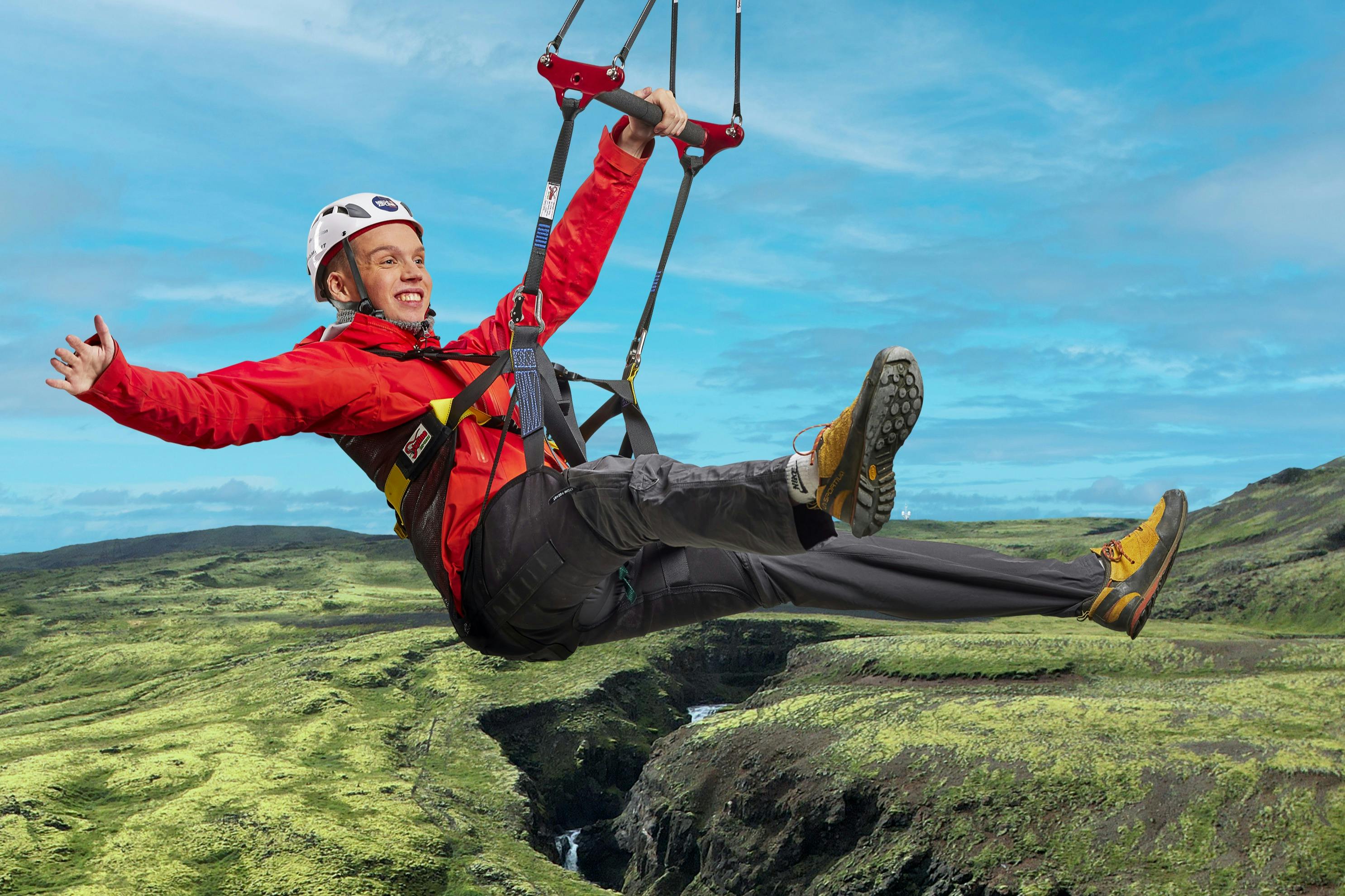 Une personne vêtue d'une veste rouge et d'un casque fait de la tyrolienne au-dessus d'un paysage de collines vertes avec une rivière en contrebas, en souriant et en écartant les bras.