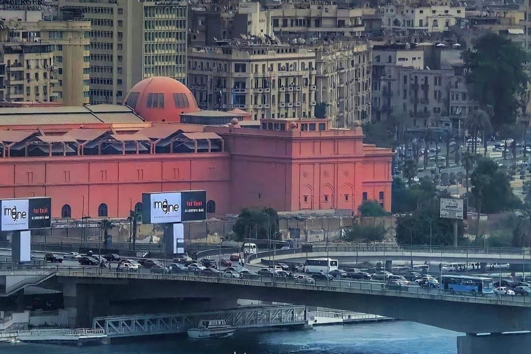 位于市区的一座大型橙色建筑和繁忙的桥梁，交通繁忙，背景是高大的米色建筑。